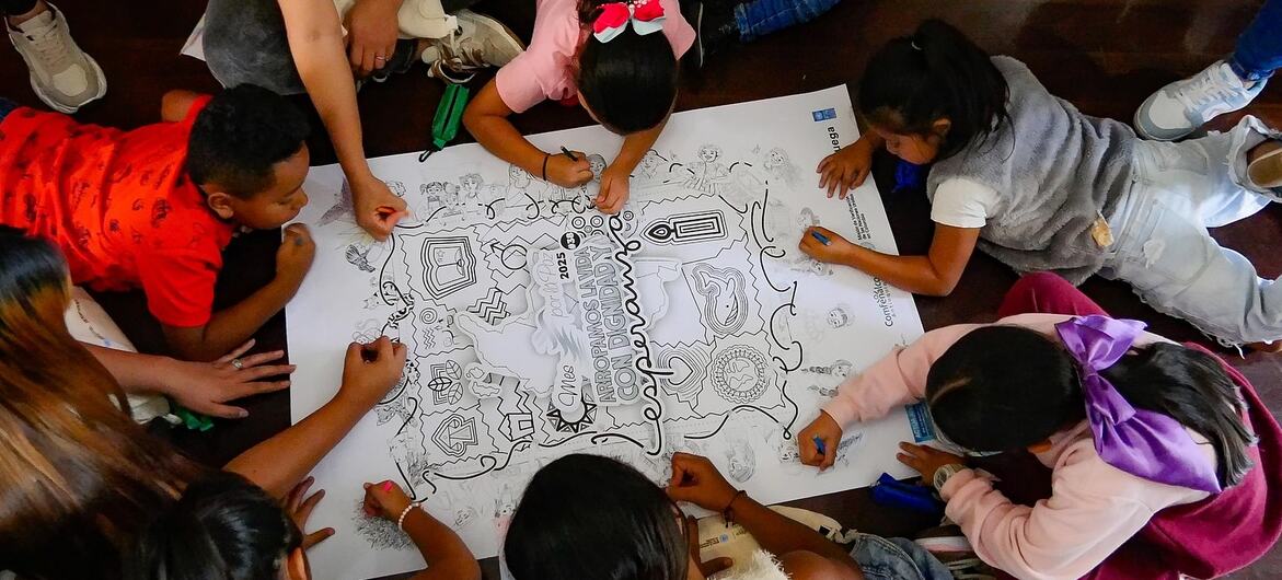 Overhead view of several children and an adult woman sitting on a wooden floor, coloring a large white UN poster with line drawings and Spanish text.