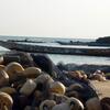 Une plage côtière de sable fin en Afrique de l'Ouest, avec des filets de pêche et des bouées au premier plan et des pirogues en bois flottant près du rivage sous un ciel brumeux.