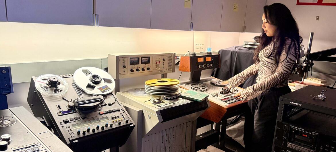 A woman in a zebra-print top operates reel-to-reel tape machines and rack-mounted audio equipment in a preservation studio.