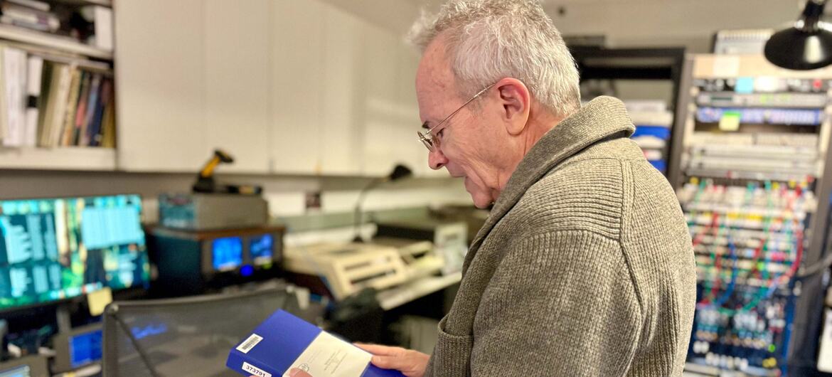 An older man with gray hair and glasses, wearing a gray ribbed sweater, holds and examines a blue archival storage box in a room lined with shelves of film reels and computer monitors.