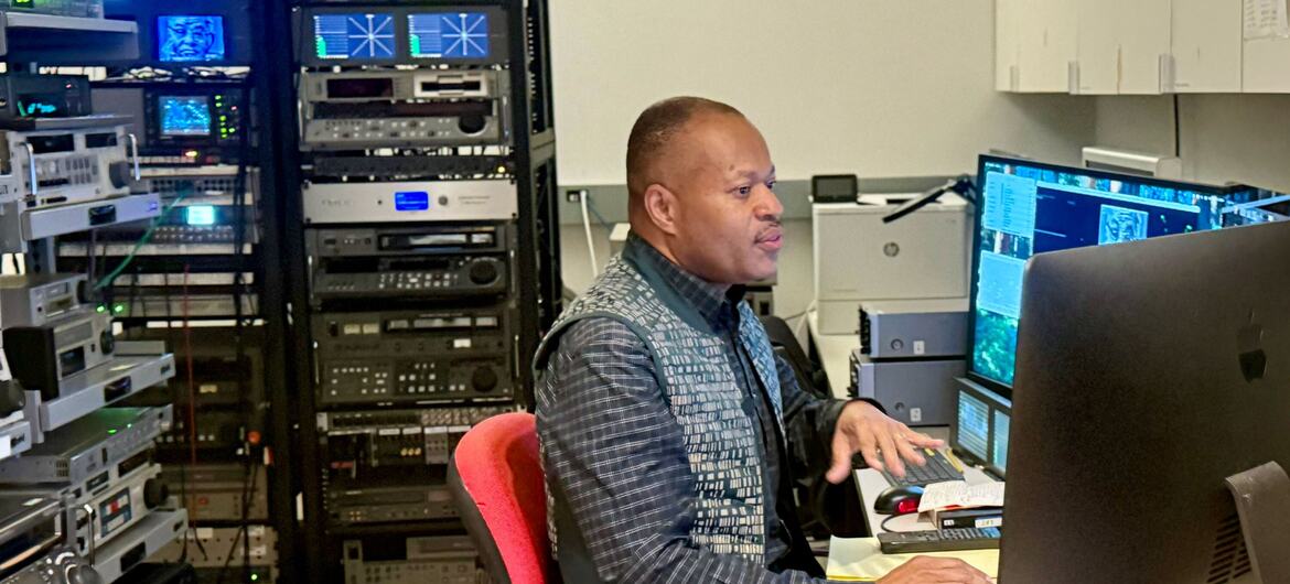 A Black male broadcast engineer works at a multi-monitor workstation in a technical control room, surrounded by racks of audio-video equipment and video monitoring screens.