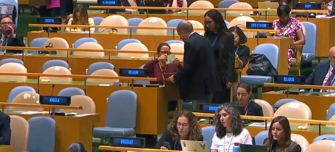 Delegados e funcionários trabalham no salão da Assembleia Geral da ONU , onde placas com os nomes de países como Bélgica, Angola, Uruguai e Bielorrússia são visíveis.