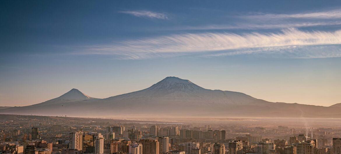 Aerial panorama of Yerevan, Armenia, with dense residential buildings in the foreground and snow-capped Mount Ararat rising behind under a clear sky.