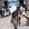 رجل يحمل كيساً أبيض كبيراً تابعاً لبرنامج الأغذية العالمي United Nations على كتفه خارج منشأة إغاثة في اليمن، بينما يصطف الناس بالقرب من لافتة للمطالبة بمساعدات طارئة للسكان النازحين.