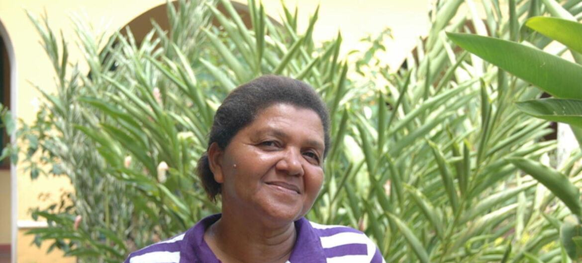 Joana Rodrigues Mousinho, uma senhora brasileira mais velha, está em um jardim ao ar livre vestindo uma camisa polo listrada em roxo e branco.