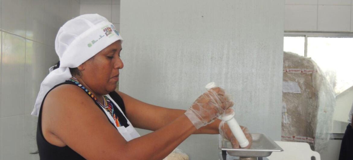 Uma mulher indígena usando uma touca de cabelo branca, blusa preta, avental branco e luvas transparentes opera um moedor de alimentos manual em uma cozinha revestida de azulejos, com sacos de grãos, bolas de massa e bandejas de ingredientes por perto.