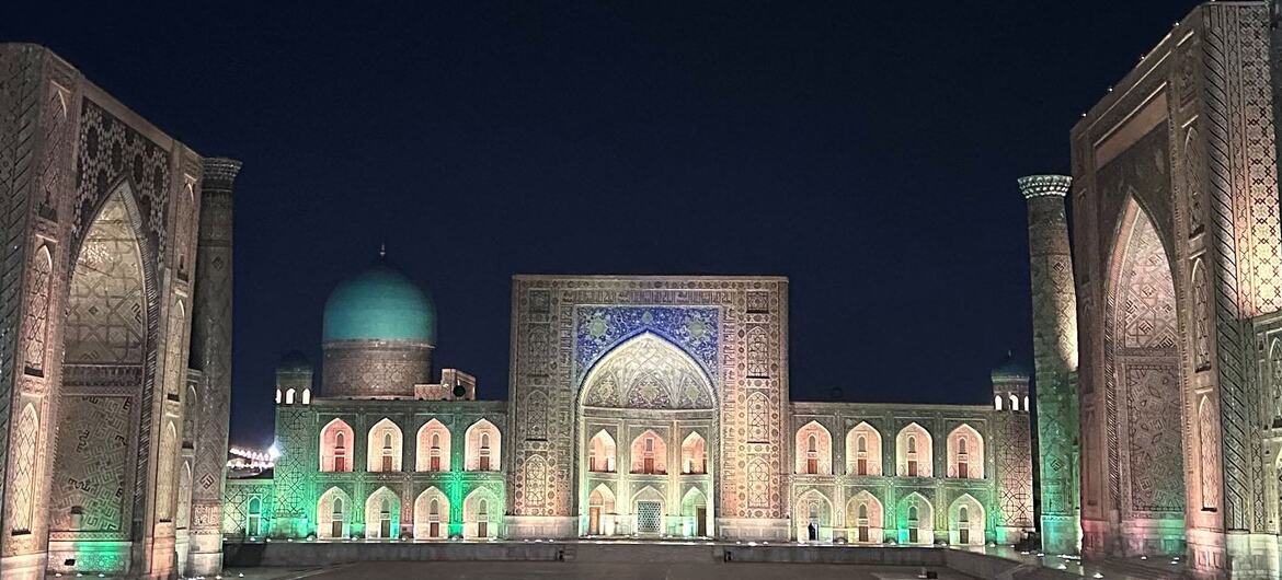 Registan square at night with illuminated historic madrasahs, minarets, a turquoise dome, and visitors gathered in the open plaza.