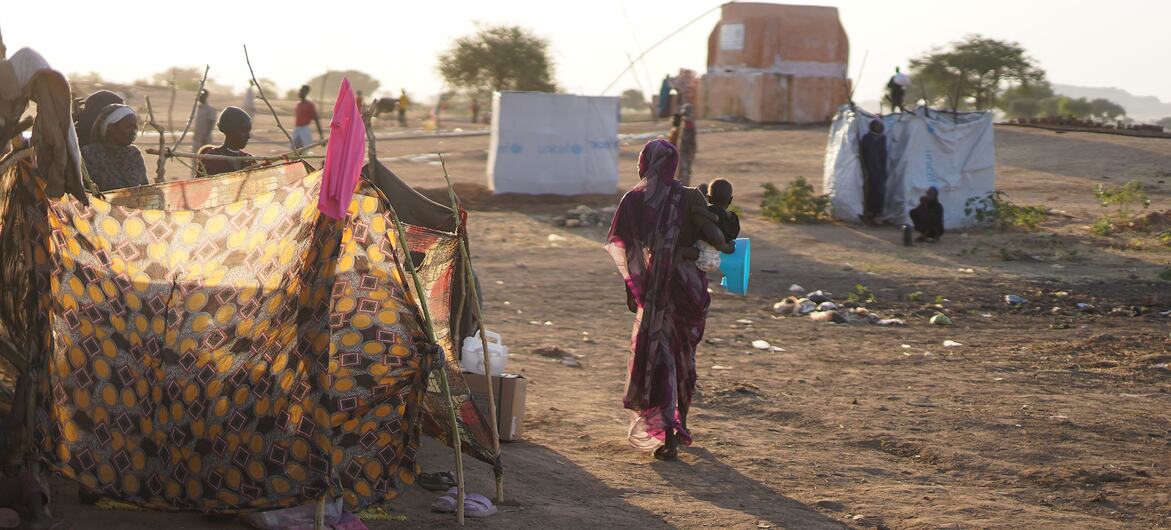 A mother walks through a displaced persons camp in Tawila, North Darfur, Sudan, carrying a child past makeshift fabric shelters and UNICEF-branded tarpaulins on dry ground.