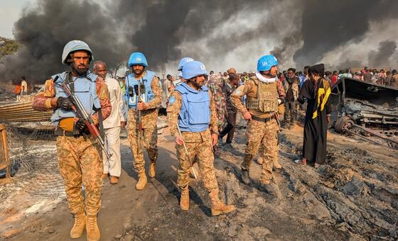 نیلے ہیلمٹ اور صحرائی کیموفلاج میں مسلح United Nations فوجی جلی ہوئی گاڑی کے پاس سے گزر رہے ہیں جب کہ گھنا سیاہ دھواں اٹھ رہا ہے، قریب ہی شہری جمع ہیں۔