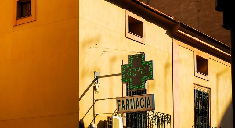 Un letrero LED verde en forma de cruz sobre un letrero de FARMACIA en un edificio amarillo indica 42 °C bajo la luz del sol, con un semáforo verde a la derecha.