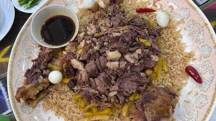 A large ornate plate of Uzbek plov with brown rice, shredded meat, yellow carrot strips, quail eggs, red chili peppers, and a small bowl of dark dipping sauce.