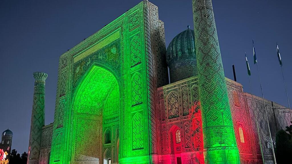 A historic Islamic architectural complex in Samarkand is illuminated with green and red lighting at night, showing a large arched portal, ornate tilework, a turquoise dome, and flagpoles against a dark blue sky.