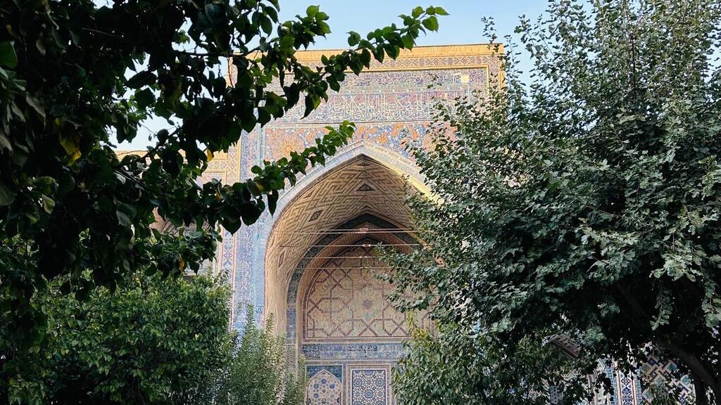 Ornate tiled Islamic archway with geometric patterns and calligraphy framed by green tree branches in Samarkand.