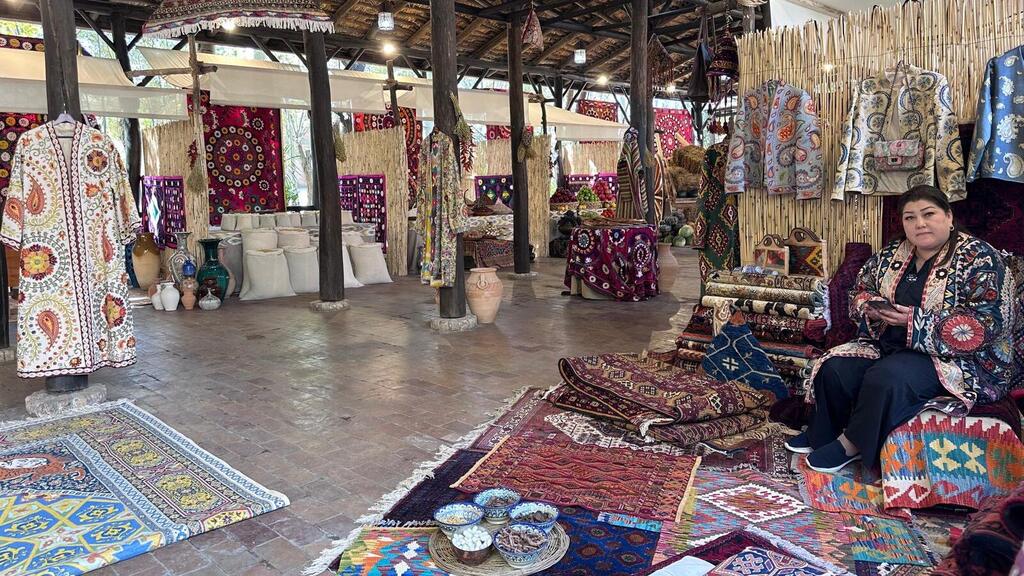 A woman sits in a traditional Samarkand craft stall surrounded by colorful carpets, embroidered dresses, cushions, ceramics, and straw-paneled walls.