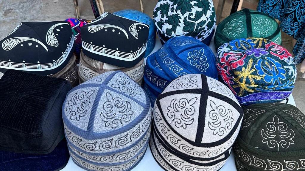 Colorful traditional embroidered felt hats displayed in stacks at a market stall, featuring geometric and floral patterns in black, blue, green, white, and multicolored designs.