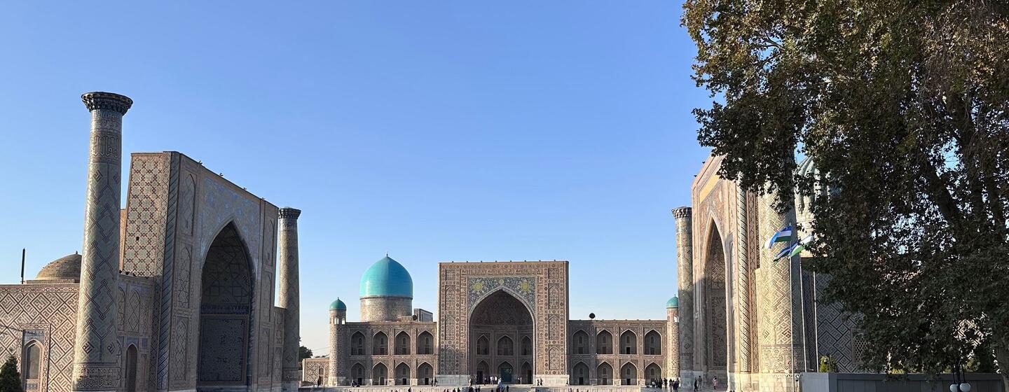 Visitors walk and sit on the stone plaza of Registan in Uzbekistan, framed by tiled madrasahs, minarets, and a turquoise dome under a clear blue sky.