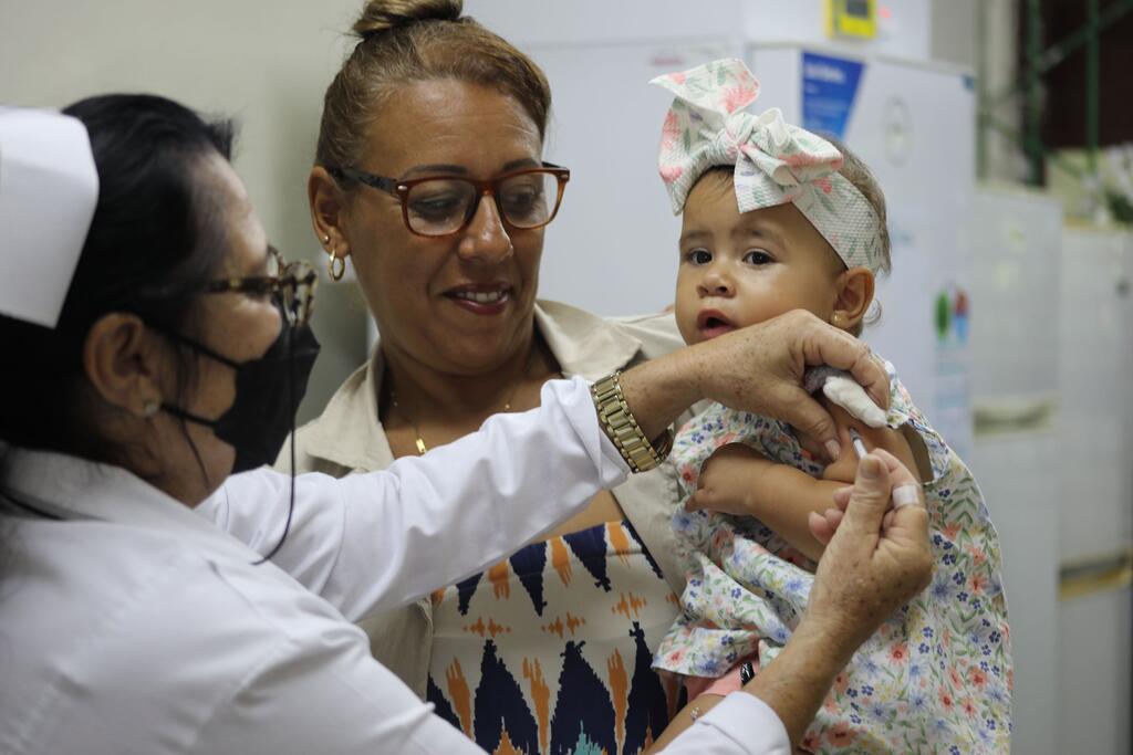 Uma enfermeira administra uma vacinação a um bebê em mãos de uma mulher, mostrando o trabalho de saúde da ONU em Cuba.