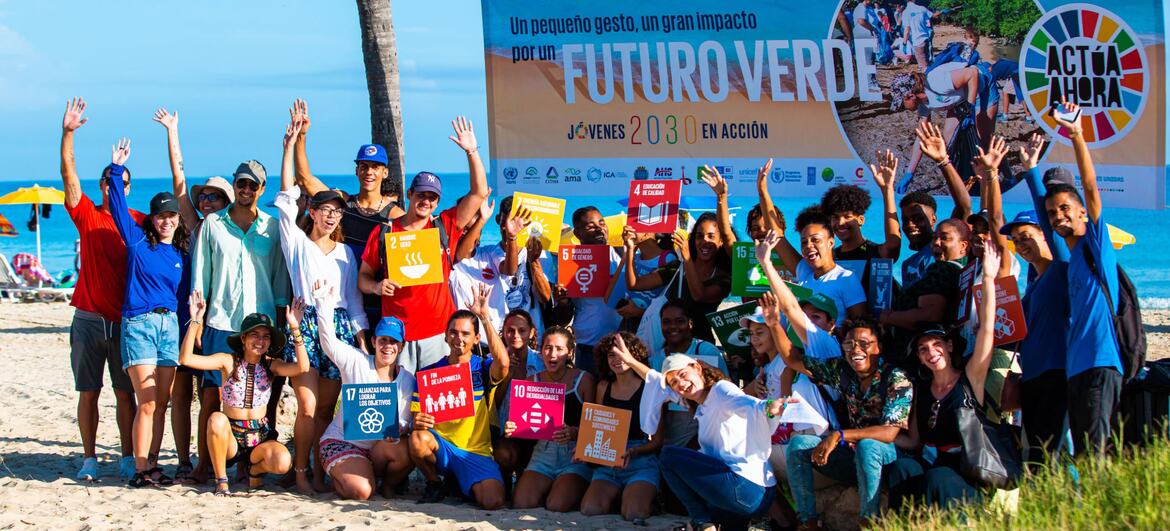 Un grupo de jóvenes en una playa sosteniendo pancartas que representan los Objetivos de Desarrollo Sostenible de las Naciones Unidas, frente a un estandarte que dice "FUTURO VERDE" y "JÓVENES 2030 EN ACCIÓN".