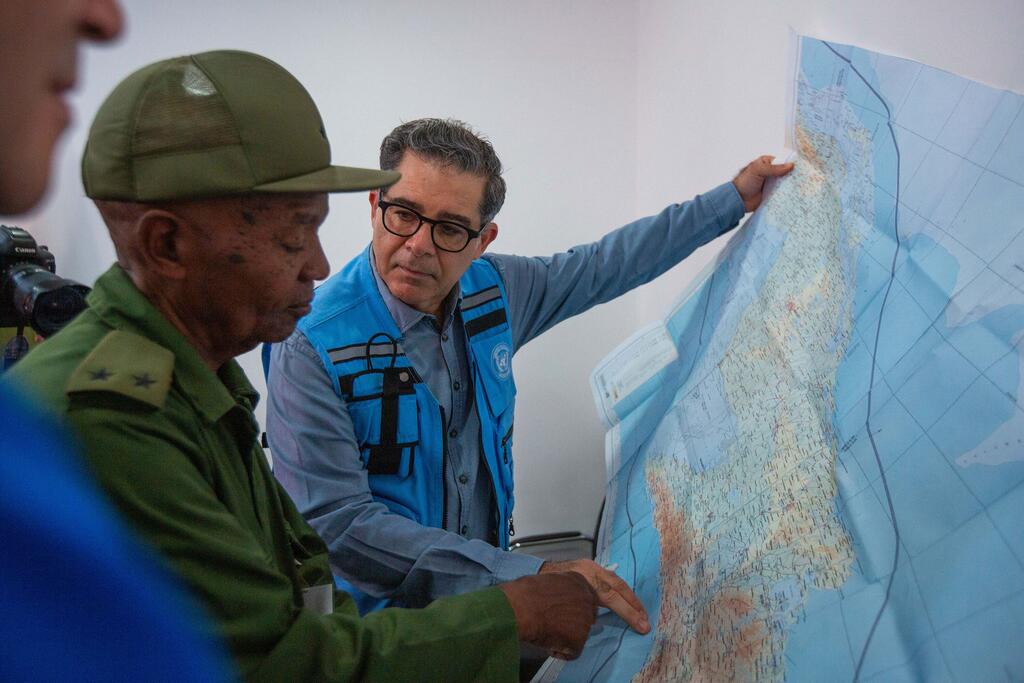 El coordinador residente de la ONU, Francisco Pichon, vestido con un chaleco azul de la ONU, señala un mapa de Cuba mientras discute con un funcionario local en uniforme militar.