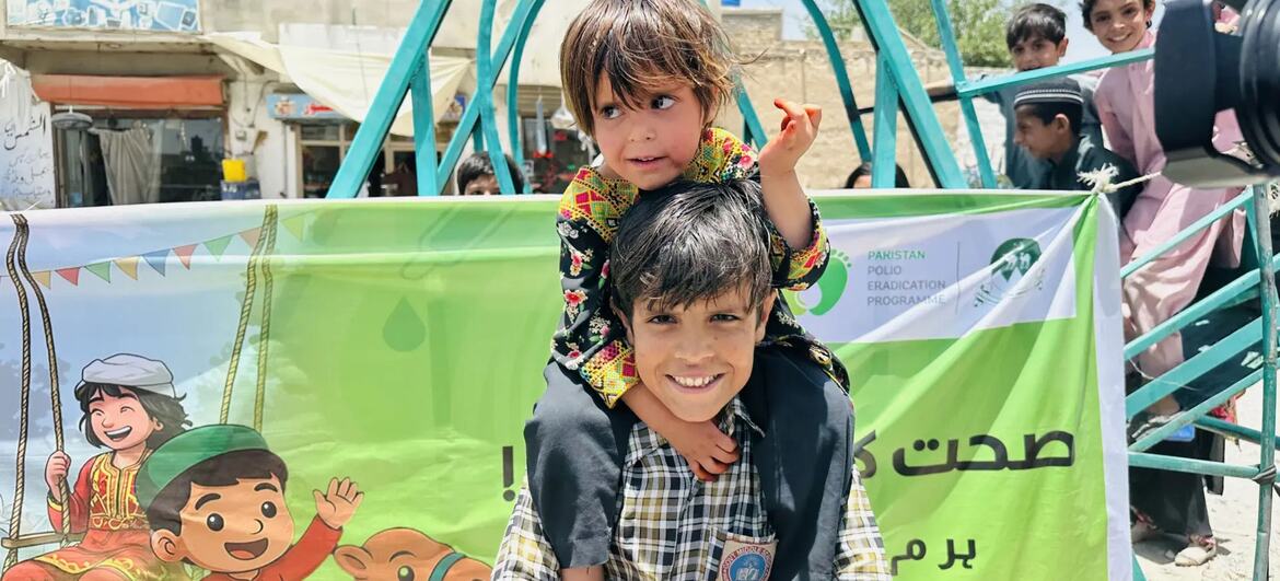 ایک مسکراتے ہوئے لڑکا پاکستان پولیو کے خاتمے کے پروگرام کے گرین بینر کے سامنے اپنے کندھوں پر ایک چھوٹا بچہ اٹھا رہا ہے۔ بینر میں بچوں کی کارٹون اور اردو میں متن دکھایا گیا ہے، جس میں پولیو ویکسینیشن کی کوششوں کو فروغ دیا گیا ہے۔