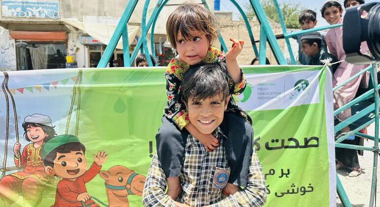 ایک مسکراتے ہوئے لڑکا پاکستان پولیو کے خاتمے کے پروگرام کے گرین بینر کے سامنے اپنے کندھوں پر ایک چھوٹا بچہ اٹھا رہا ہے۔ بینر میں بچوں کی کارٹون اور اردو میں متن دکھایا گیا ہے، جس میں پولیو ویکسینیشن کی کوششوں کو فروغ دیا گیا ہے۔