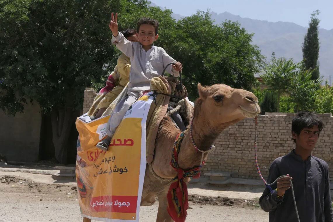 بلوچستان، پاکستان میں یونیسیف اور جی آئی سی اے کی حمایت سے پولیو ویکسینیشن مہم کے دوران ایک اونٹ پر مسکراتے ہوئے ایک بچے نے امن کے اشارے کو ہلایا۔ ایک مقامی لڑکا اونٹ کی قیادت کرتا ہے۔ اونٹ پر پولیو ویکسینیشن کی تشہیر کرنے والا ایک بینر ہے۔