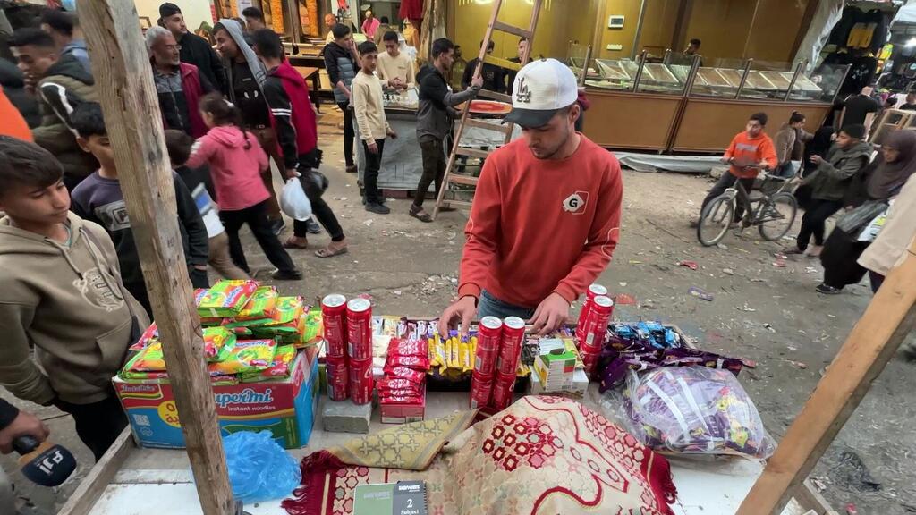 A young Palestinian man in a red sweatshirt and white cap sells snacks and candy from a makeshift stall in a crowded market in Khan Younis, Gaza.