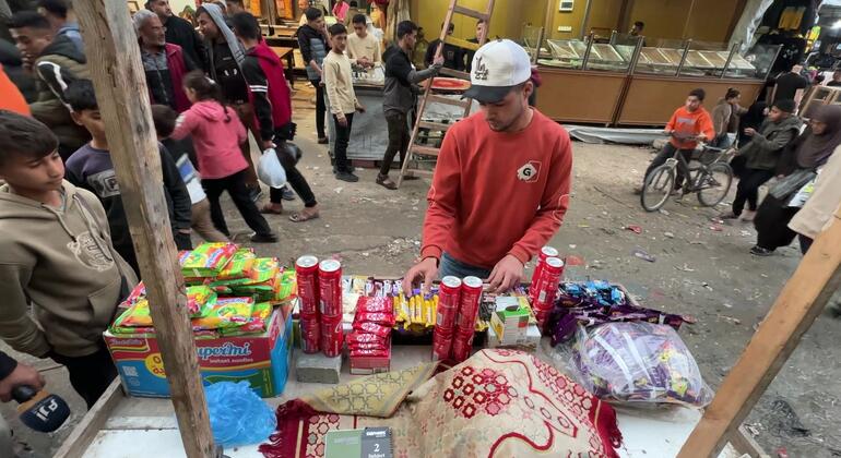 A young Palestinian man in a red sweatshirt and white cap sells snacks and candy from a makeshift stall in a crowded market in Khan Younis, Gaza.