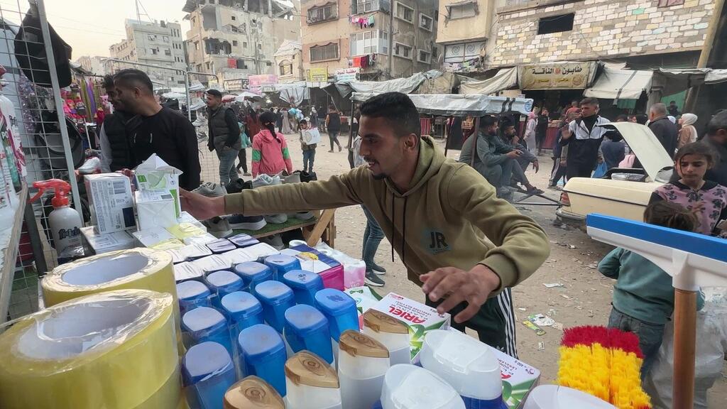 A Palestinian man sells household goods like cleaning supplies and tape at a makeshift street market in Khan Younis, Gaza. In the background, damaged buildings are visible, illustrating the 'survival economy' where educated youth take on simple jobs due to the ongoing conflict.
