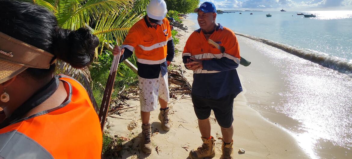 Construction workers wearing high-visibility orange vests and hard hats on a sandy beach lined with coconut trees prepare for coastal protection works in Funafuti, Tuvalu.