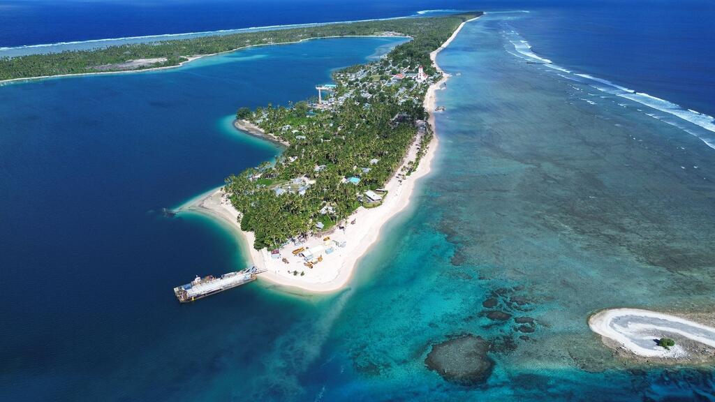 Aerial view of the island state of Tuvalu showing its narrow strip of land surrounded by a bright turquoise lagoon and a deep blue ocean, highlighting the effects of coastal erosion and climate change.