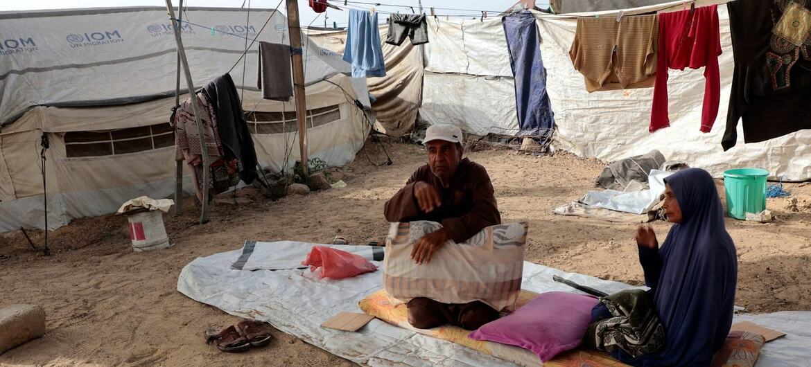 رجل وامرأة يجلسان على حصيرة في مخيم للاجئين في غزة الرملي، مع خيام المنظمة الدولية للهجرة وغسيل الملابس معلقة في الخلفية.