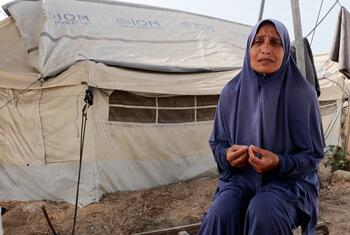 A woman in a blue headscarf sits in front of a white tent with IOM branding in a refugee camp.