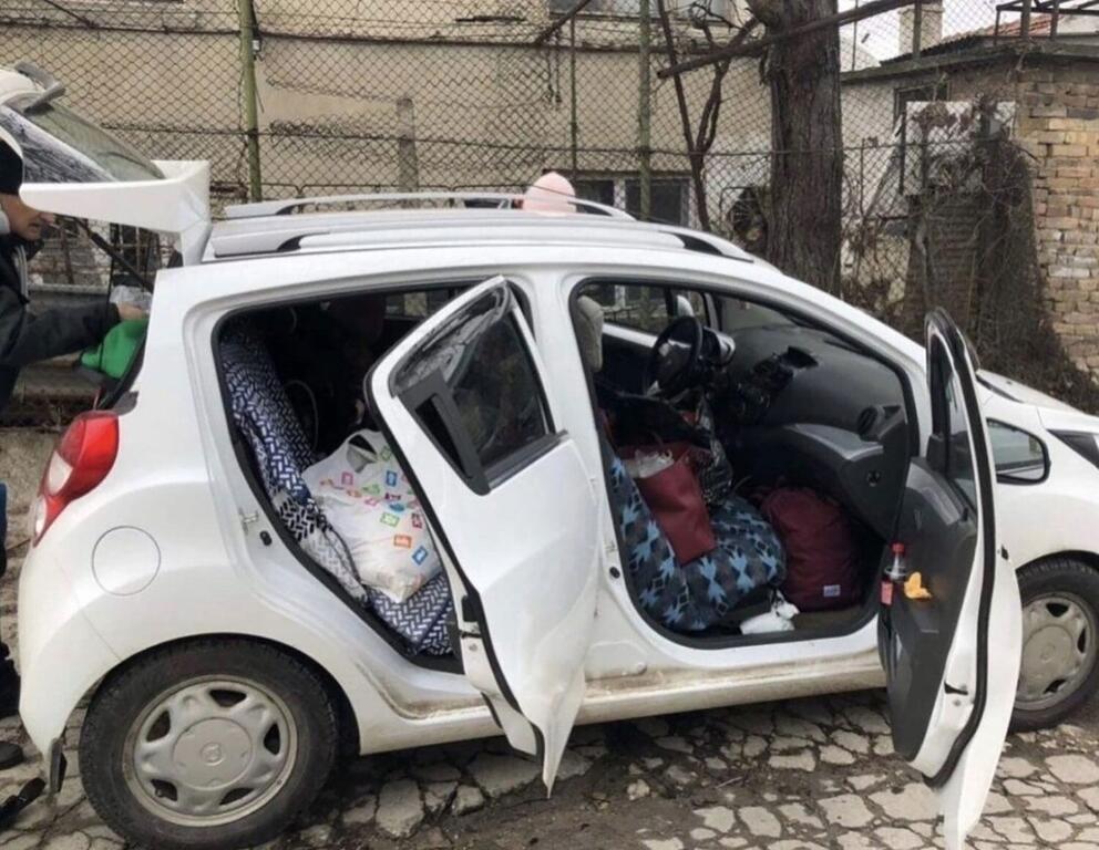 Белый хэтчбек загружен вещами и багажом, двери открыты. Сзади автомобиля виден человек, который, по-видимому, загружает или выгружает предметы.