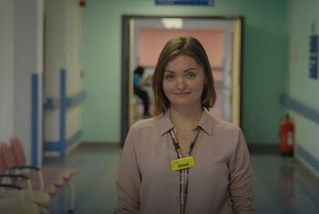 A Ukrainian woman, Dr Inna Soldatenko, stands in a hospital corridor in the UK. She is wearing a lanyard with an ID badge that reads 'Inna'.