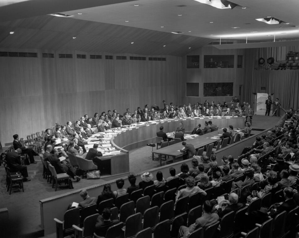 Черно-белая фотография первого заседания Второй сессии Экономического и Социального Совета Организации Объединенных Наций, на которой делегаты из разных стран сидят вокруг большого изогнутого стола с аудиторией на переднем плане.