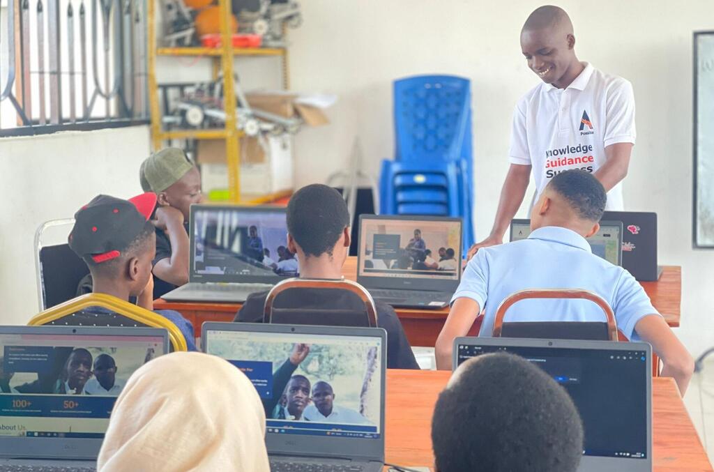 Wanafunzi nchini Tanzania wanashiriki katika darasa la elimu ya ICT, wakitumia kompyuta ndogo na wakiongozwa na mwalimu katika mazingira ya maabara ya kompyuta.
