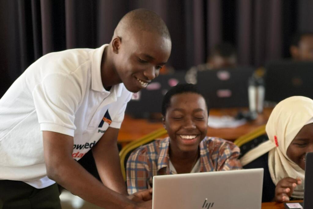 Mwanamume mchanga mwenye tabasamu akiwa amevaa shati nyeupe la polo anasimama ili kusaidia wanafunzi wawili wa kike wanaotabasamu wanapofanya kazi kwenye kompyuta ndogo darasani.