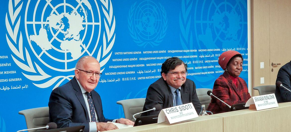Tres personas sentadas en una mesa de conferencia de prensa de la ONU con placas de nombres, en un telón de fondo con el logotipo de la ONU y el texto multilingüe.