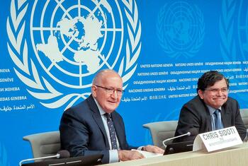 ثلاثة أشخاص جالسون على طاولة مؤتمر صحفي للأمم المتحدة مع لوحة أسماء، في الخلفية مع شعار الأمم المتحدة والنص متعدد اللغات.