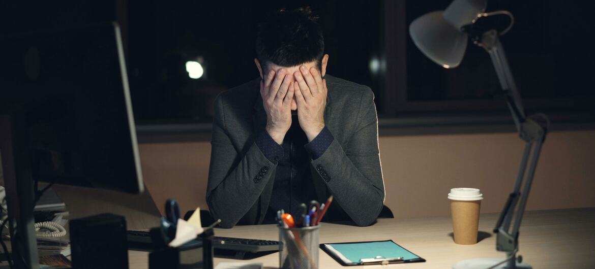 A tired young man in a suit sits at a dark office desk at night, holding his head in his hands in front of a computer screen, illustrating stress and burnout.