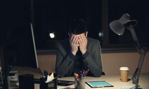 A tired young man in a suit sits at a dark office desk at night, holding his head in his hands in front of a computer screen, illustrating stress and burnout.