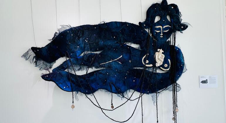 Большая синяя скульптура из волокна, изображающая лежащую фигуру со спокойным лицом, украшенную бусинами и ниспадающими кисточками, экспонируемая на белой стене на выставке искусства коренных народов.
