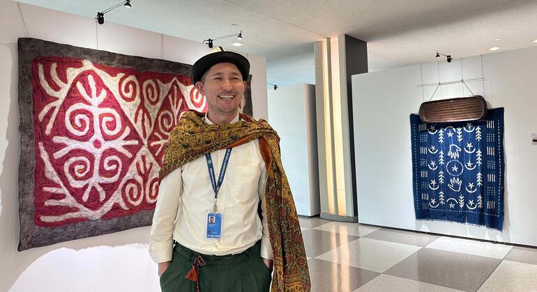 Улыбающийся мужчина стоит в выставочной галерее, одетый в чёрную шляпу, кремовую рубашку и узорчатый платок. За ним на стене висят два больших текстильных произведения искусства коренных народов: одно красное с белыми спиральными узорами, другое — синее с белыми символами.