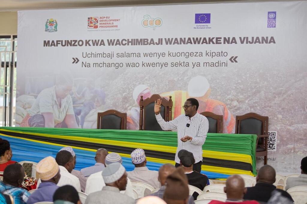 Mwanamume aliyevaa shati lenye mistari anawasilisha hotuba kwenye mkutano kuhusu uchimbaji madini nchini Tanzania. Bendera kubwa nyuma yake inaonyesha nembo za UNDP, ACP-EU Development Minerals Programme, na Umoja wa Ulaya, pamoja na maandishi ya Kiswahili.