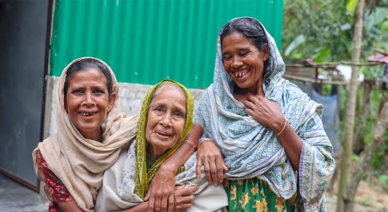 Wanawake watatu wenye tabasamu kutoka Bangladesh, pamoja na mwanamke mzee aliye katikati, wanakumbatiana katika mazingira ya vijijini.