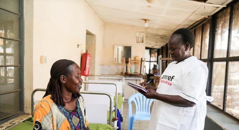 Muuguzi wa kike amevaa sare nyeupe yenye maandishi "Amref Health Africa" yaliyochapishwa, anaandika kwenye kitabu cha kumbukumbu akiwa amesimama kando ya mgonjwa wa kike ameketi katika chumba cha hospitali.