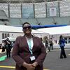 Mwanamke mweusi mwenye tabasamu akiwa amevaa suti ya kahawia na miwani ya jua anapozana Mkutano wa Kijijini wa Dunia 13 huko Baku, ambapo kuna mabango ya tukio hilo na jumba kubwa la ndani nyuma.