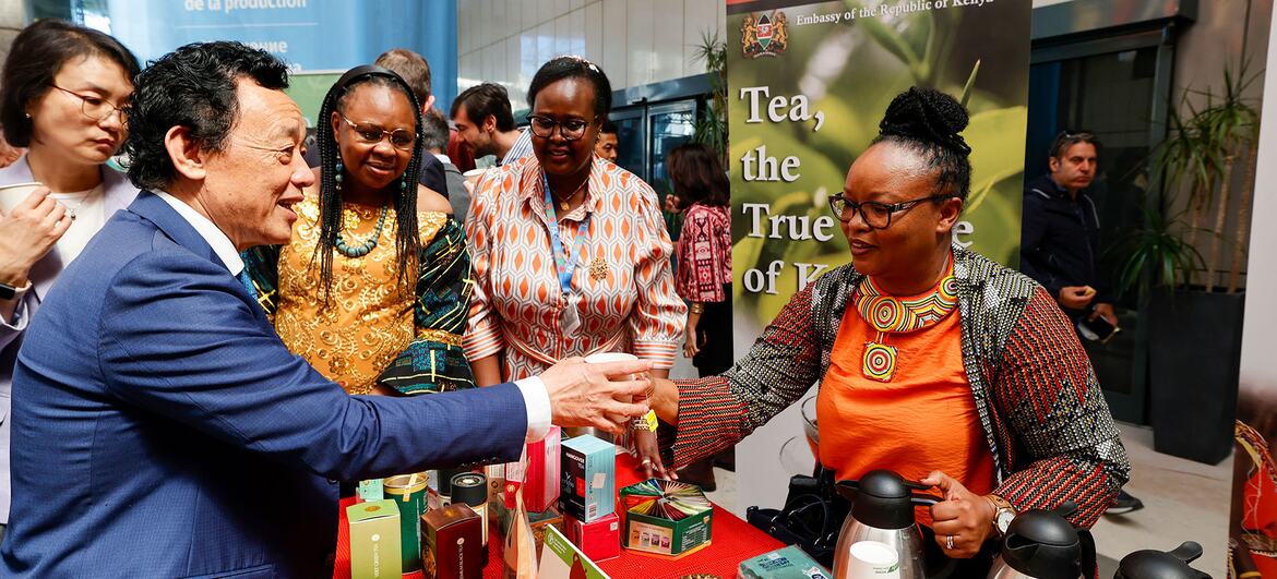 A man in a blue suit shakes hands with a woman in traditional attire at an Embassy of the Republic of Kenya booth featuring tea products.