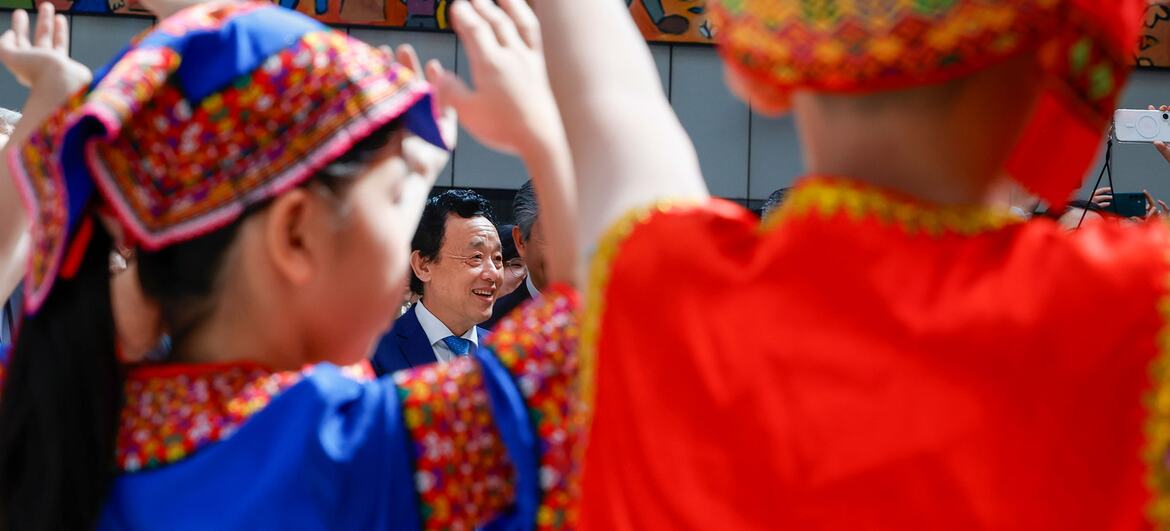 身着多彩传统服饰与帽子的中国儿童正在表演舞蹈，一位面带微笑的西装男子正与他们握手。